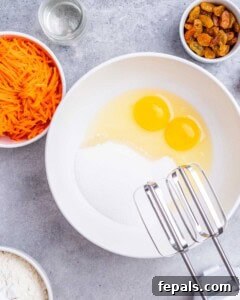 Bowl with sugar and two cracked eggs ready to be mixed with a hand mixer, surrounded by grated carrots and raisins.