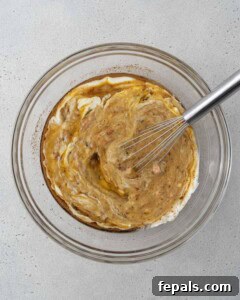 mashed banana, yogurt, egg, oil, and sugar are added to a bowl to be mixed for banana bread.
