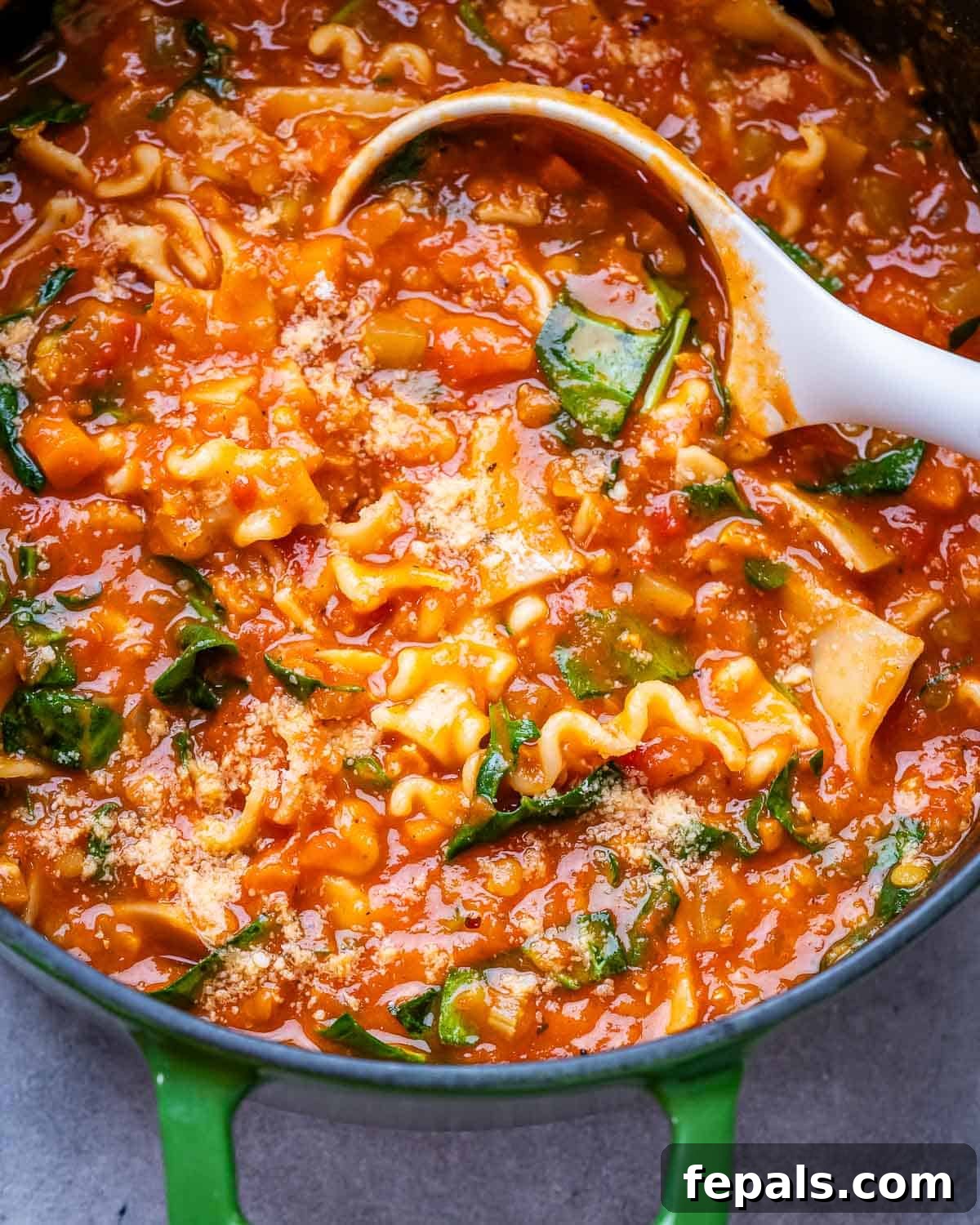 A vibrant green pot filled with vegetarian lasagna soup, featuring a white ladle.