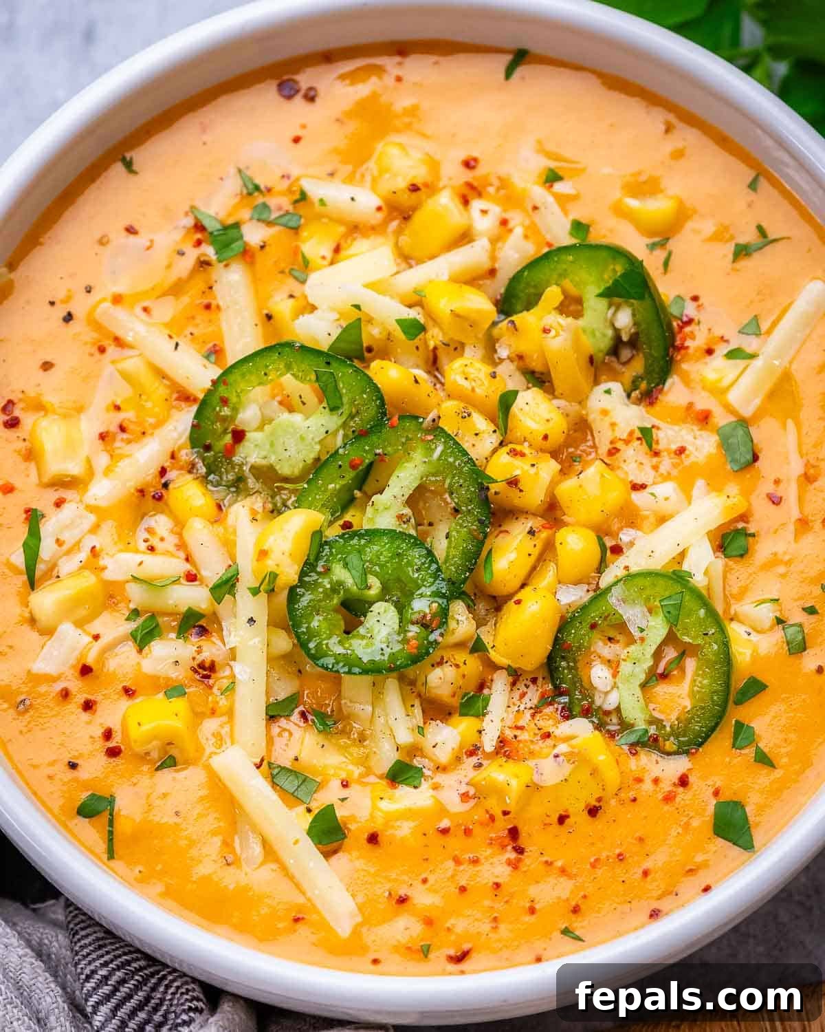 A white bowl of creamy Mexican Street Corn Soup, generously topped with crumbled queso fresco, fresh cilantro, and a few slices of vibrant green jalapeño. The soup has a rich, golden hue, suggesting its corn base and the warmth of the spices.