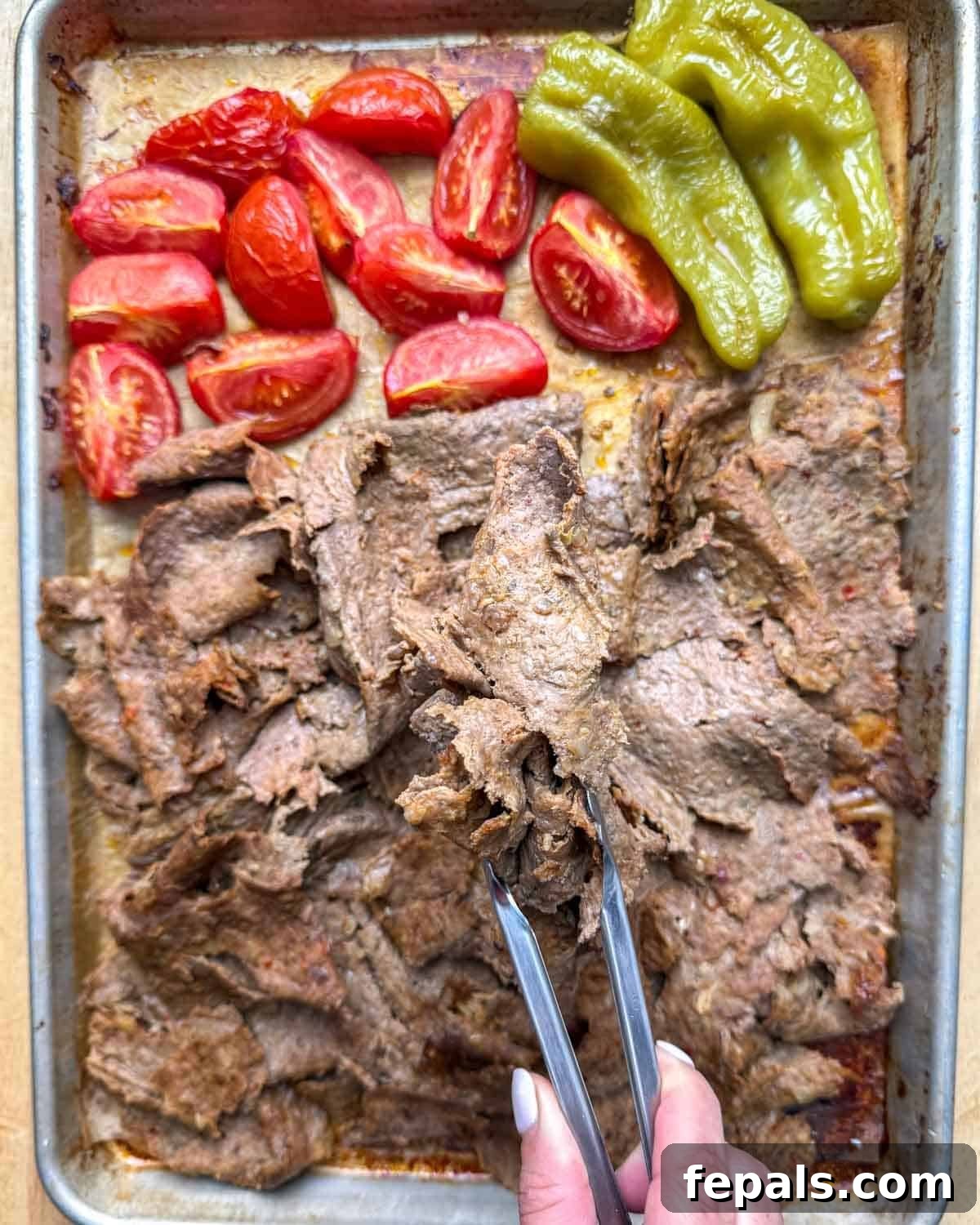 Kitchen tongs picking up a handful of freshly shredded doner kebab from a baking pan, highlighting its crispy texture.