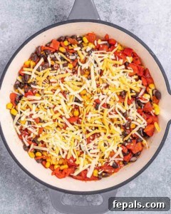 Overhead view of a skillet filled with shredded cheese, corn, black beans, and red peppers before baking.