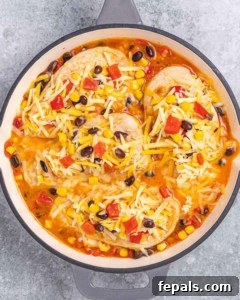 Skillet of chicken breasts covered in shredded cheese, ready to bake with a layer of beans, corn, and peppers underneath.