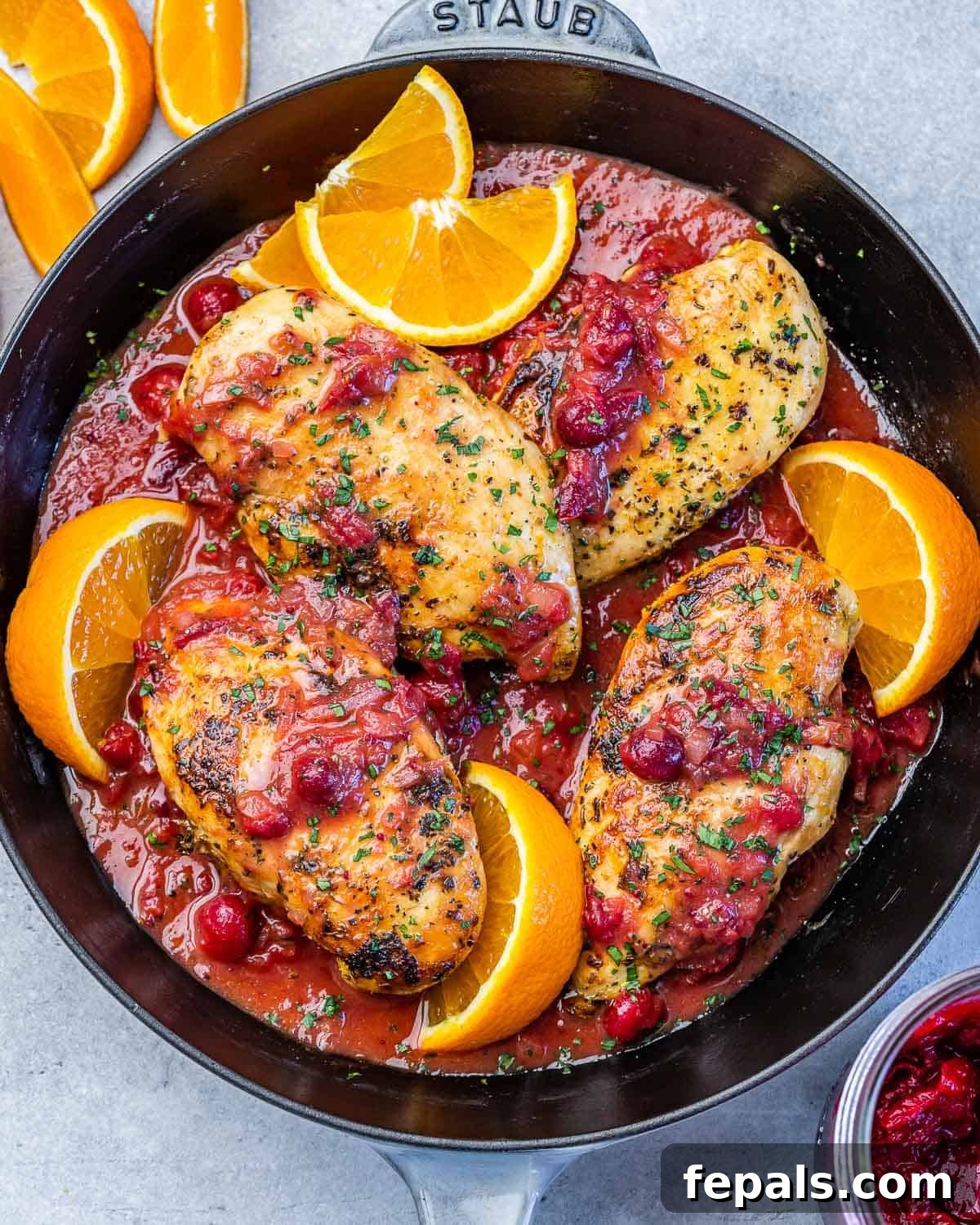 Zesty Cranberry Orange Chicken 2 Top view of a black skillet that has 4 cooked chicken breasts in an orange cranberry sauce with orange slice garnishes around the chicken.