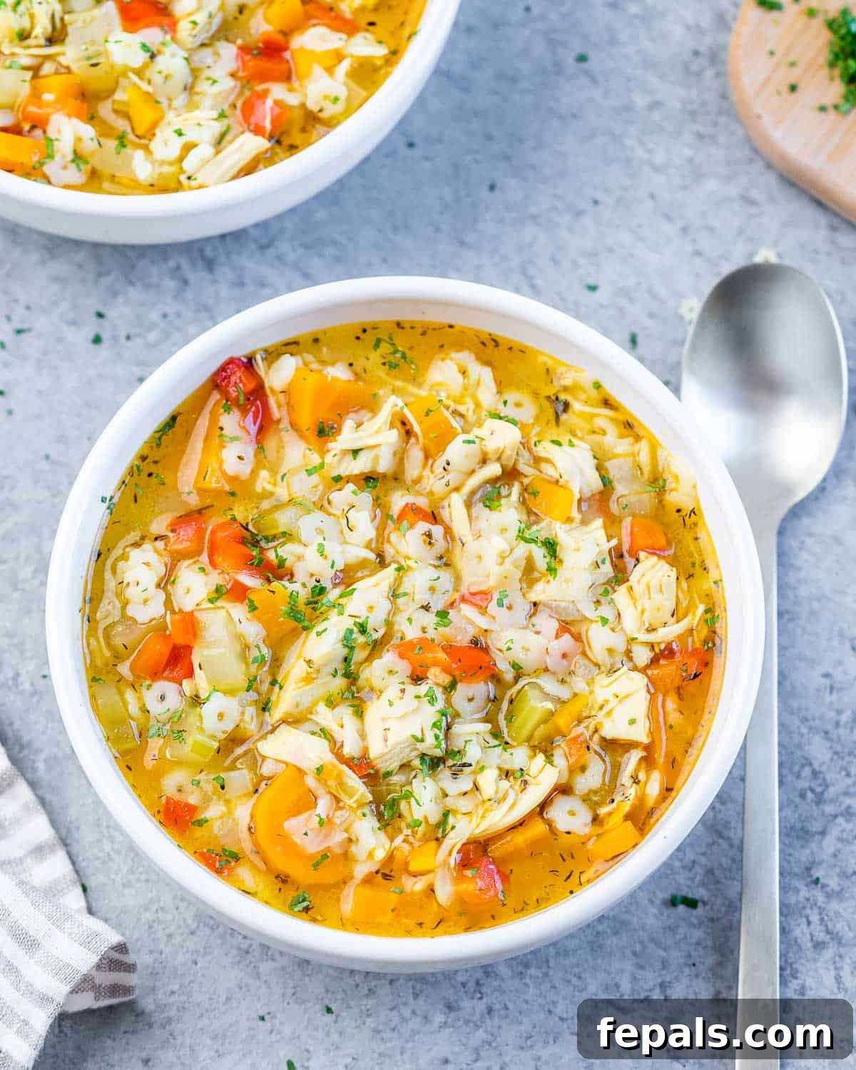 A white bowl of chicken pastina soup with a striped napkin and spoon, ready to be enjoyed.
