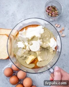 Overhead shot of ingredients in a food processor, including butter, pistachio butter, cream cheese, and honey, ready to blend for the custard base of a brioche French toast casserole.