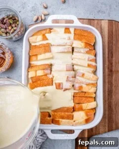 Pouring the pistachio custard over neatly layered brioche slices in a white casserole dish, preparing for baking.