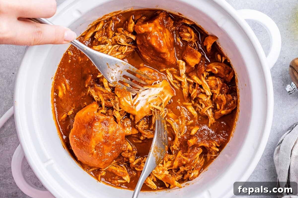Effortless Slow Cooker BBQ Pulled Chicken 2 Hand using two forks to shred BBQ chicken inside a crockpot with sauce, highlighting the tender texture.
