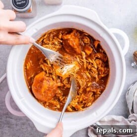 Hand shredding cooked BBQ chicken with two forks inside a white crockpot.