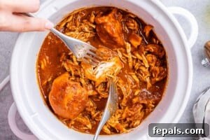 Effortless Slow Cooker BBQ Pulled Chicken 15 Hand using two forks to shred BBQ chicken inside a crockpot with sauce.