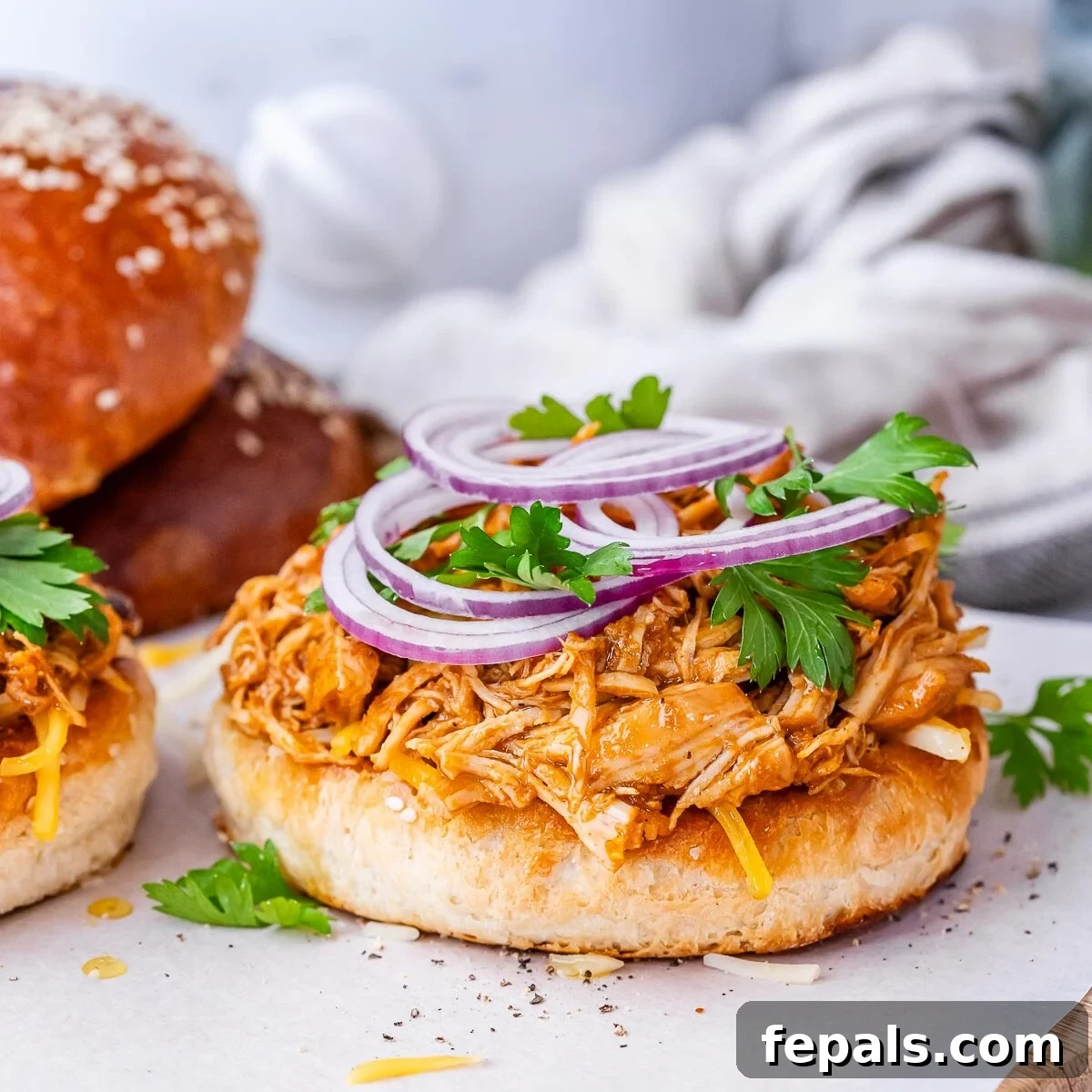 Effortless Slow Cooker BBQ Pulled Chicken 7 An open-faced bun generously loaded with homemade BBQ shredded chicken and garnished with vibrant sliced red onion, with another prepared sandwich in the background.