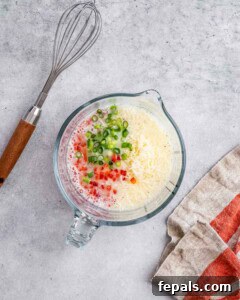 Mixing bowl with egg white mixture, red pepper, green onion, and shredded cheese, with a metal whisk resting inside.