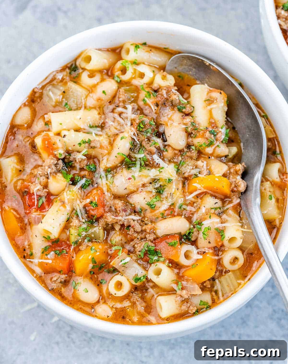 A white bowl of pasta fagioli soup with a spoon, garnished with fresh parsley.