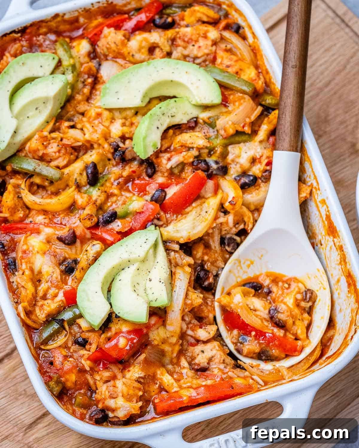 A warm, cheesy chicken fajita casserole in a white baking dish, ready to be served, with a serving spoon resting inside.