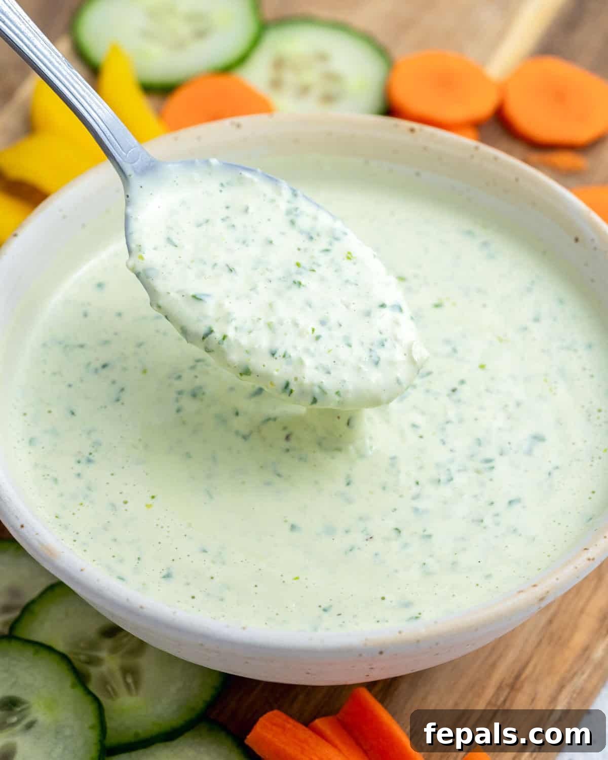 A white bowl of green goddess dressing being scooped with a spoon, surrounded by fresh vegetables. 