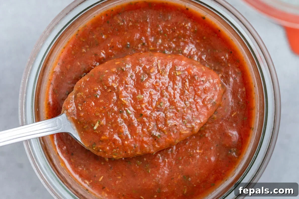 A spoonful of rich, red pizza sauce held above a clear glass jar filled with more homemade pizza sauce, ready for use.