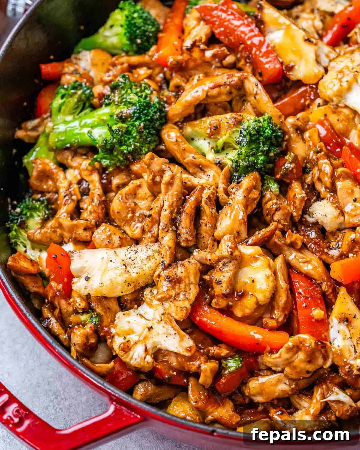 A close-up of the finished Black Pepper Chicken stir-fry in a large pan, showcasing glossy, dark sauce coating tender chicken strips, vibrant red bell peppers, fresh green broccoli florets, and white cauliflower pieces.