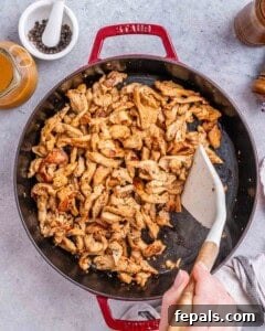 Action shot in a large red-handled pan: stir-frying marinated chicken pieces with sliced red bell peppers, broccoli florets, and cauliflower, with a white spatula tossing the ingredients.