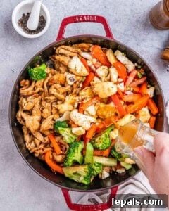 Stir-fry in progress in a large pan on a gray surface: tender chicken pieces, red bell pepper strips, broccoli, and cauliflower florets being cooked together with visible glossy sauce beginning to coat everything.