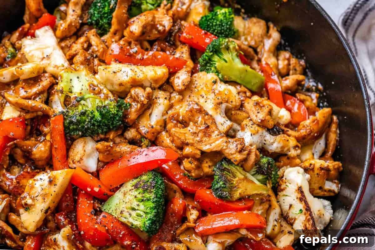Close-up shot of plated Black Pepper Chicken stir-fry, showing succulent chicken pieces, vibrant red bell pepper strips, green broccoli, and white cauliflower, all generously coated in a thick, shiny black pepper sauce.