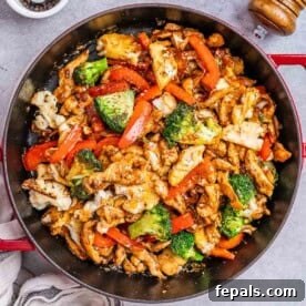 Overhead view of the completed black pepper chicken stir-fry in a red-handled pan, showing glossy sauce-glazed chicken strips mixed with bright red bell peppers, green broccoli, and white cauliflower florets.