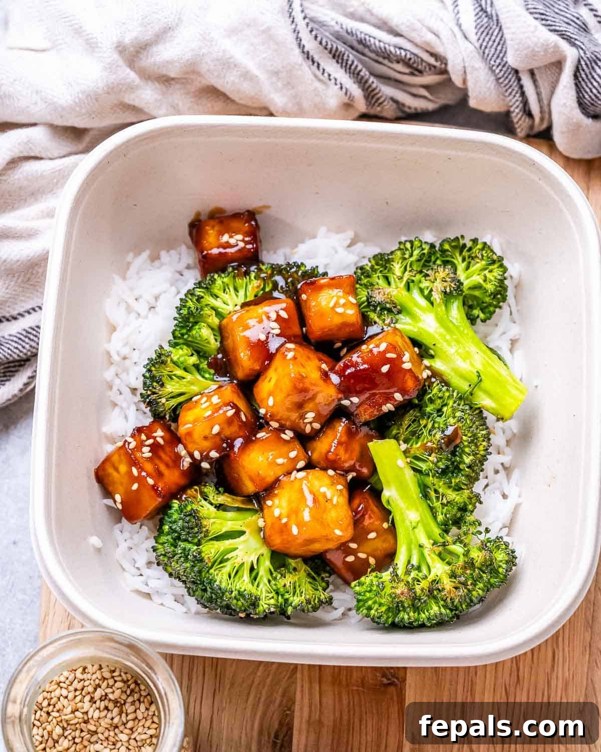 No-Fuss Teriyaki Tofu 2 A beautifully arranged white bowl featuring golden teriyaki tofu pieces served over a bed of steamy white rice, accompanied by perfectly roasted green broccoli florets. Garnished with sesame seeds.