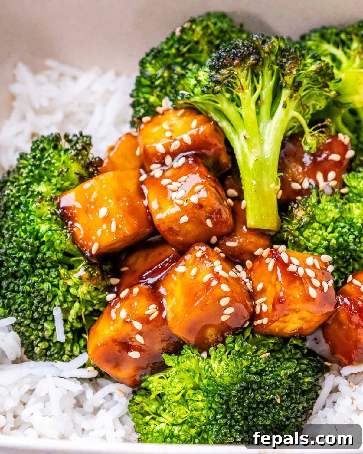 No-Fuss Teriyaki Tofu 7 A close-up shot of a bowl of white rice topped generously with baked broccoli and flavorful teriyaki tofu bites, garnished with sesame seeds.