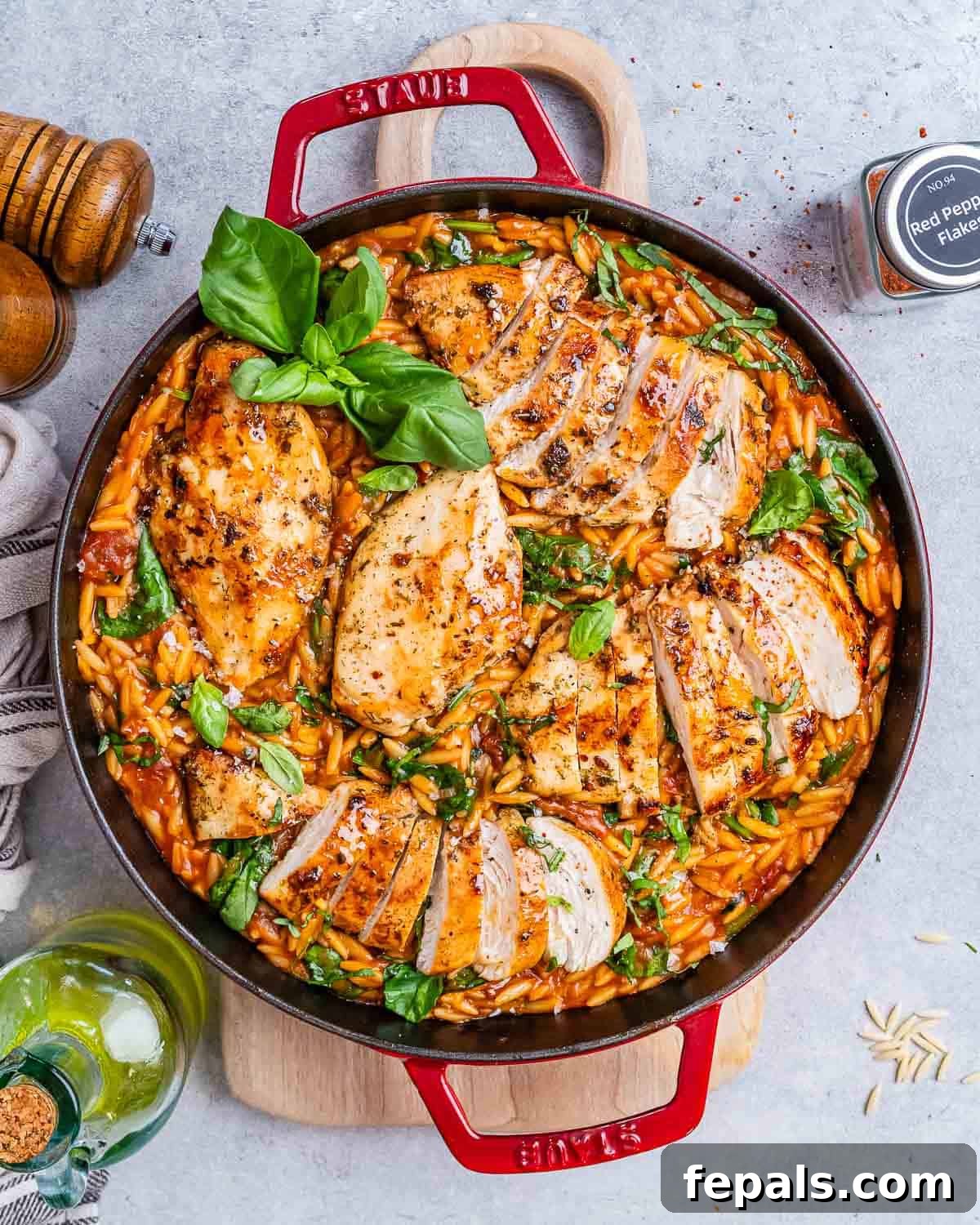 Top view of creamy Tuscan Chicken Orzo in a large red Dutch oven, showing sliced chicken on top of orzo mixed with wilted spinach, sun-dried tomatoes, and fresh basil garnish.