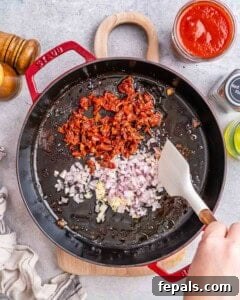Close-up of sautéing chopped sun-dried tomatoes, diced shallots, minced garlic, and red pepper flakes in oil inside a black skillet.