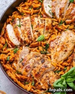 Top view of the completed dish in a red Dutch oven with juicy sliced Tuscan chicken breasts resting over vibrant orzo pasta mixed with spinach, sun-dried tomatoes, and fresh basil.