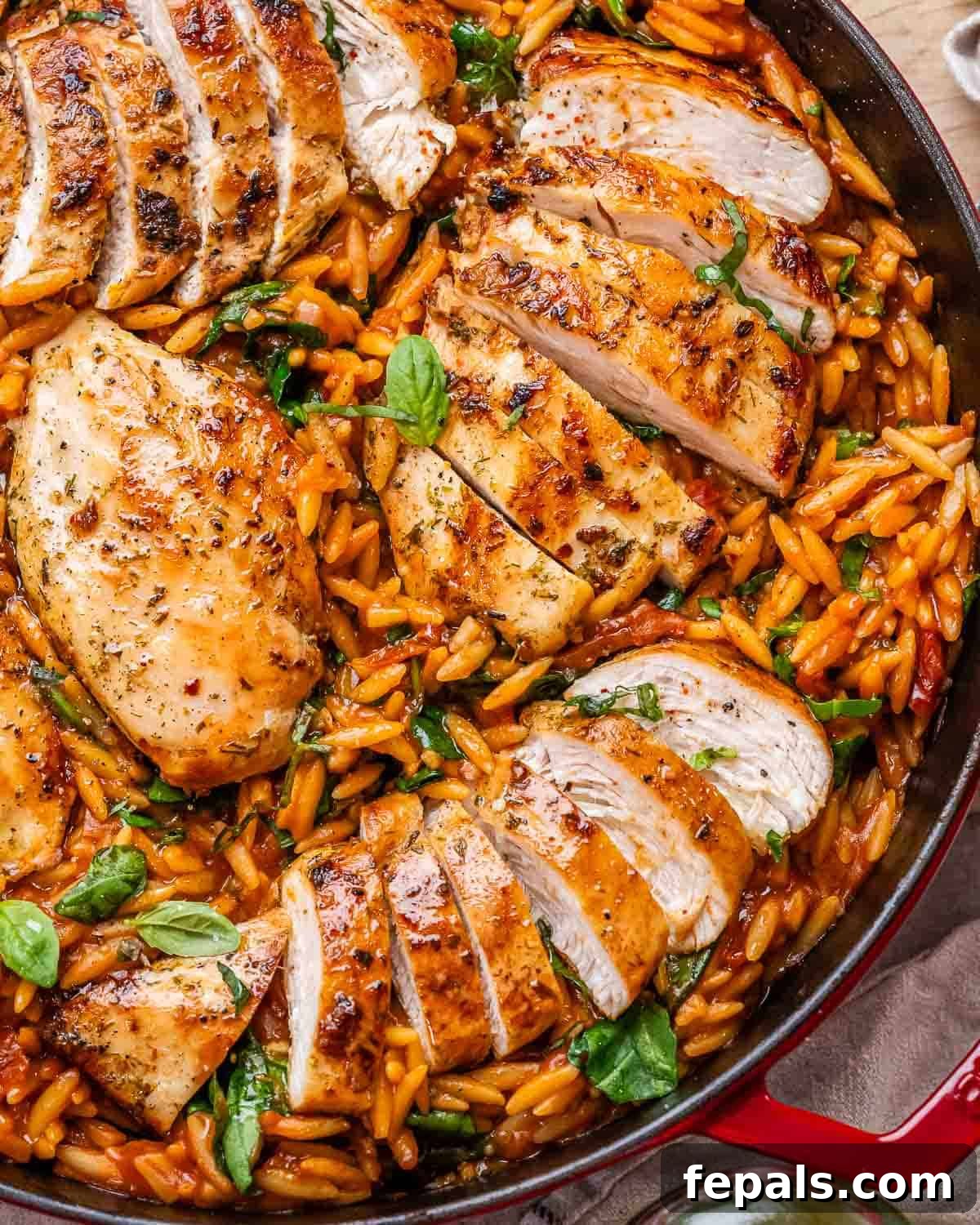 Close-up of the completed dish in a red Dutch oven with juicy sliced Tuscan chicken breasts resting over vibrant orzo pasta mixed with spinach, sun-dried tomatoes, and fresh basil.