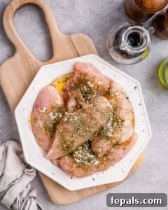 Seasoned raw chicken breasts coated in Italian seasoning and garlic powder, resting on a white octagonal plate with a wooden board of fresh basil and spinach in the background.