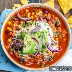 A rustic bowl of red chicken chili, garnished generously with shredded cheese, creamy avocado slices, and a dollop of sour cream.