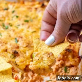 A hand scooping creamy Buffalo chicken dip with a tortilla chip from a festive bowl, showing off its cheesy texture.