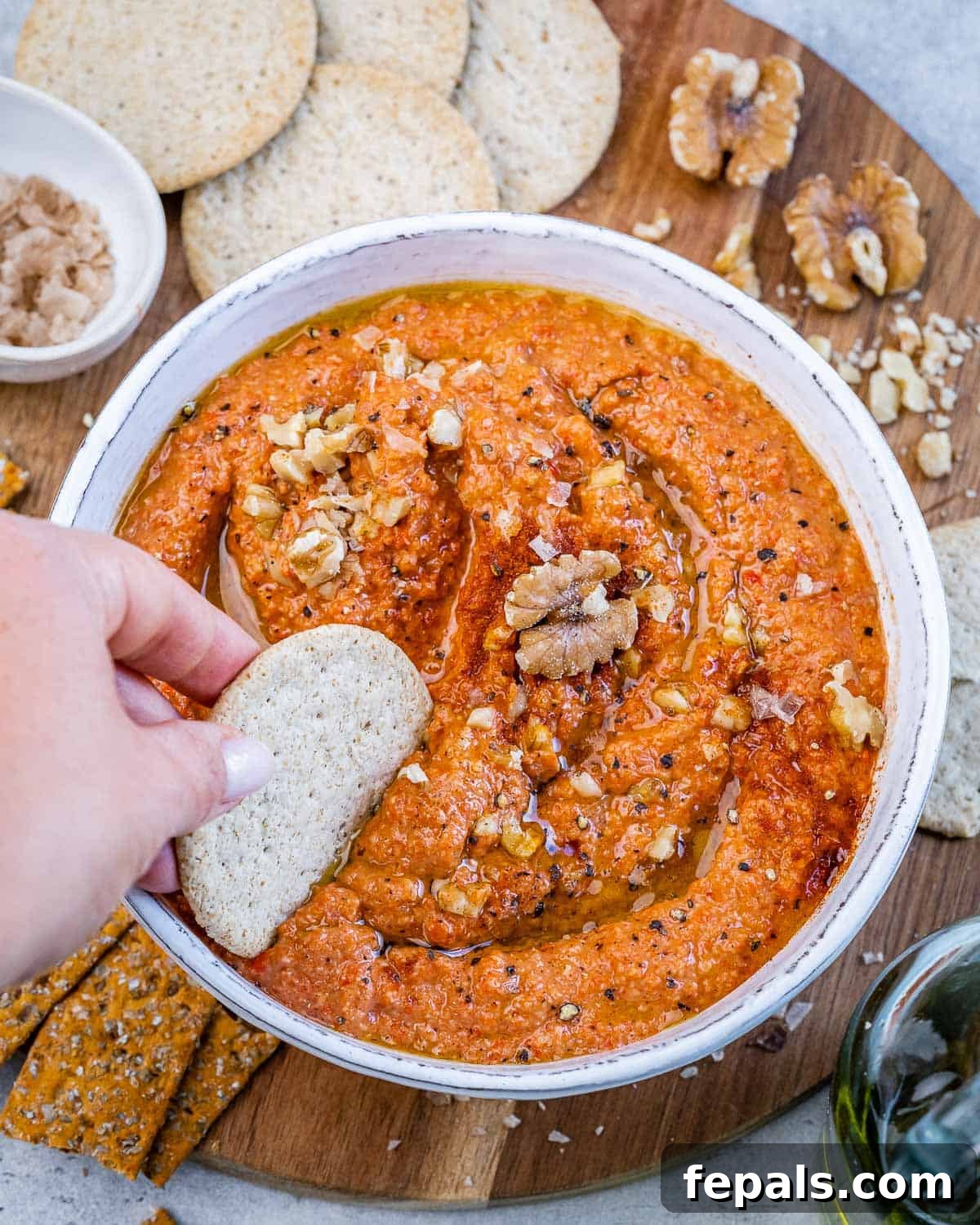 Hand dipping a pita chip into a bowl of vibrant red muhammara dip, garnished with olive oil and a sprinkle of chopped walnuts.