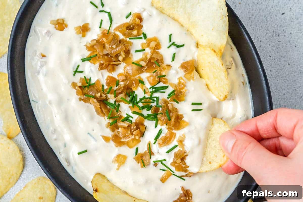 Decadent French Onion Dip 8 A hand dipping a potato chip into a bowl of creamy French onion dip, set against a bright, inviting background.