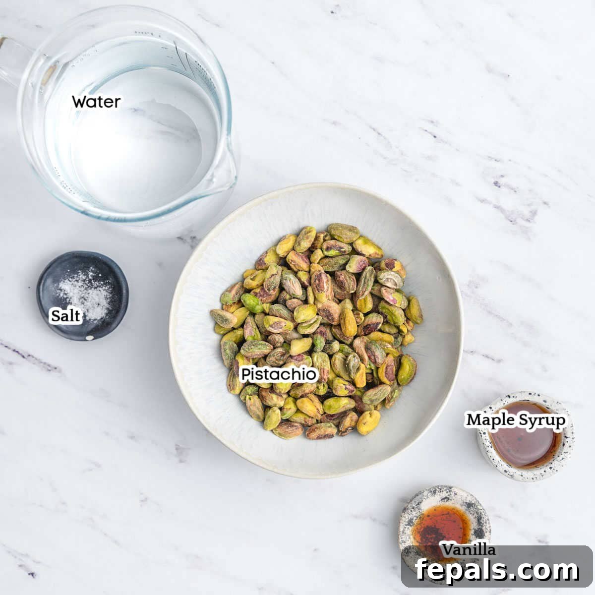 A collection of ingredients laid out, including shelled pistachios, a bottle of maple syrup, and vanilla extract.
