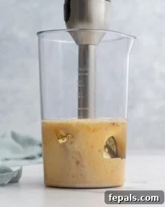 Immersion blender lifted slightly, showing more of the mixture emulsifying into a pale yellow, creamy consistency.