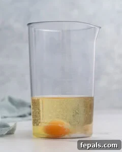 All ingredients combined in a tall glass jar before blending: whole egg at the bottom, Dijon mustard, lemon juice, vinegar, salt, and avocado oil poured on top.