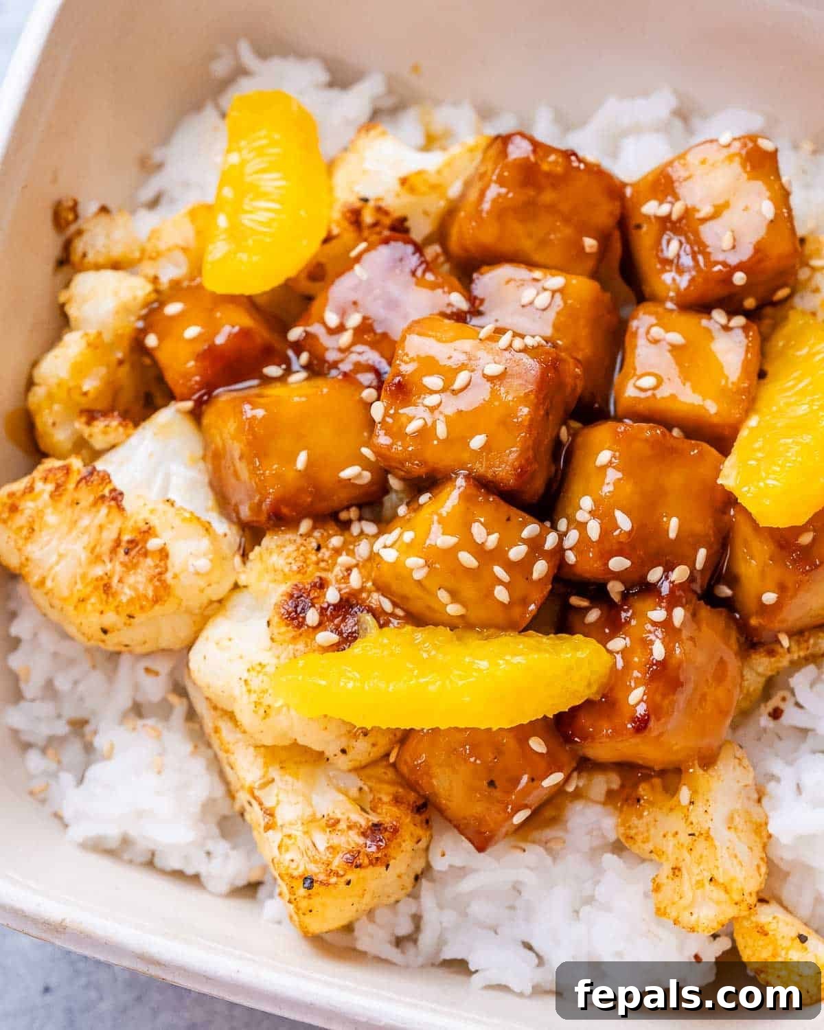 Crispy Orange Tofu 2 A close-up image of crispy orange tofu and cauliflower with rice in a white bowl, garnished with sesame seeds.