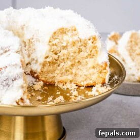 Coconut bundt cake on a gold cake stand, sliced open to reveal a soft, moist crumb and snowy coconut topping.