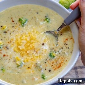 A hand holding a spoon in a bowl of healthy Broccoli Cheddar Soup, beautifully topped with a generous sprinkle of shredded cheese.