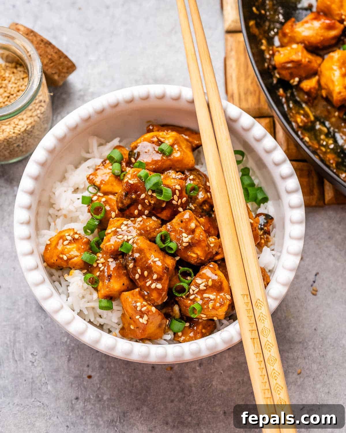 A white bowl with Hoisin Chicken and rice, garnished with scallions and sesame seeds, with chopsticks on the side. The chicken pieces are glazed in a dark, glossy sauce.