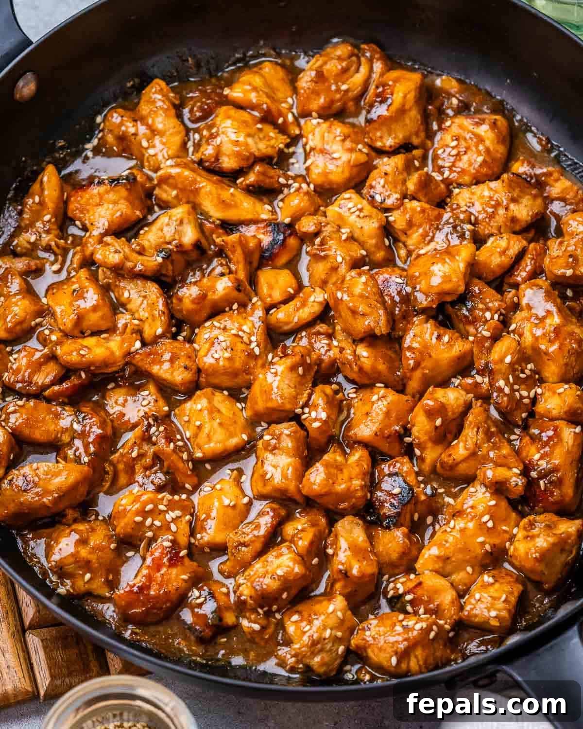 A skillet filled with glistening hoisin chicken breast chunks, generously garnished with white sesame seeds and a few green onion slices.