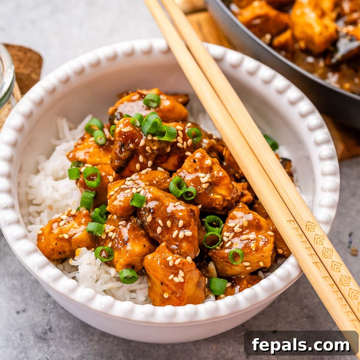 A beautifully presented bowl of Hoisin Chicken served over a bed of white rice, garnished with fresh green onions and sesame seeds. Chopsticks rest beside the bowl.