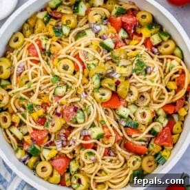 A round white bowl showcasing cold spaghetti salad, generously mixed with an assortment of fresh vegetables and savory olives.