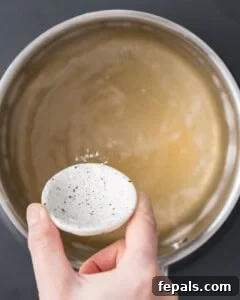 Ruby Red Pickled Eggs 12 Large stainless steel pot containing warm golden-brown pickling brine with a hand adding in ingredients from a shallow dish.