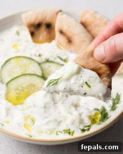 A triangle of pita bread dipped in Greek tzatziki dip held above a bowl of the dip.
