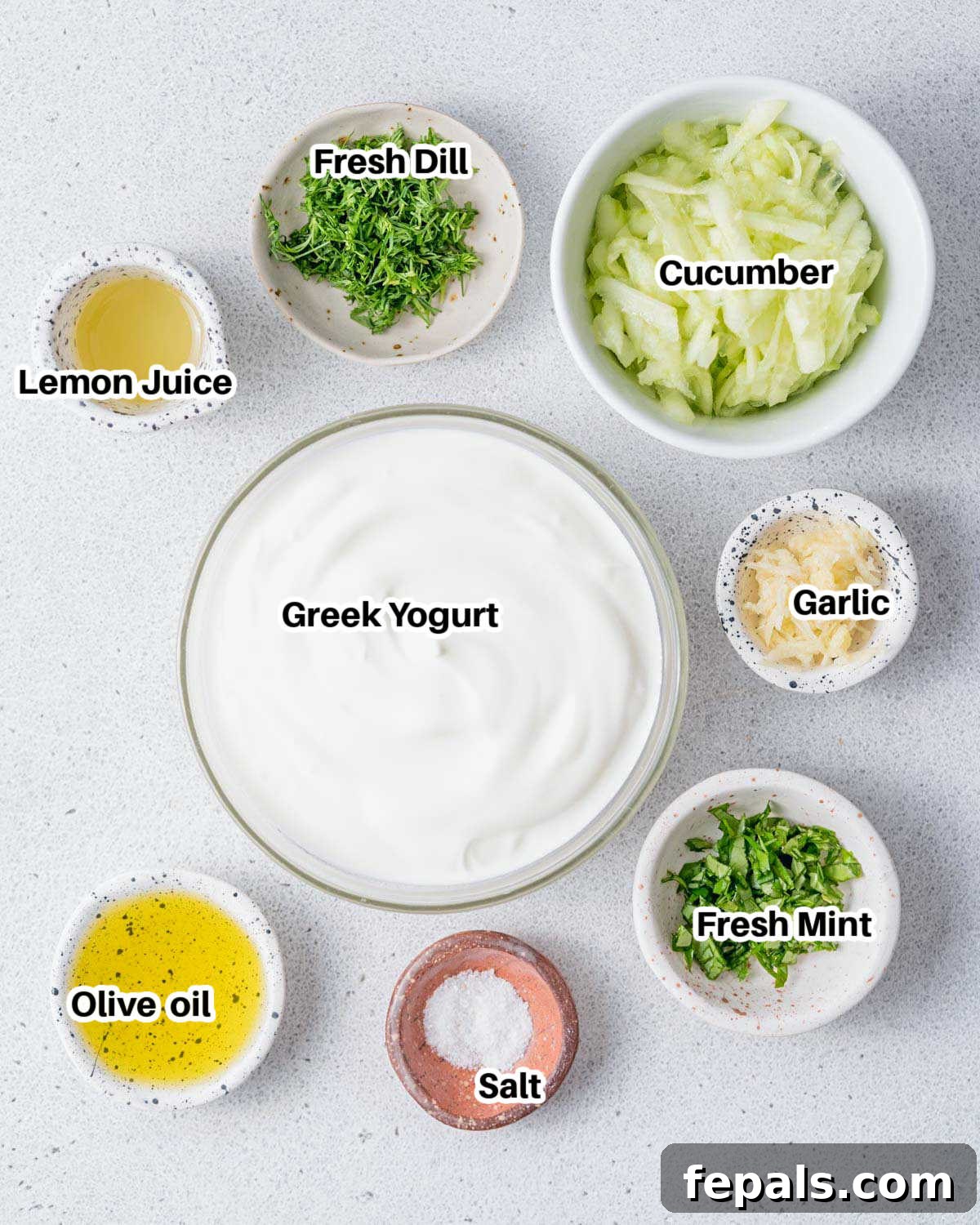 A collection of fresh ingredients laid out on a table, including a bowl of Greek yogurt, fresh cucumbers, a lemon, garlic cloves, a bottle of olive oil, and bunches of fresh dill and mint, ready to make tzatziki sauce.