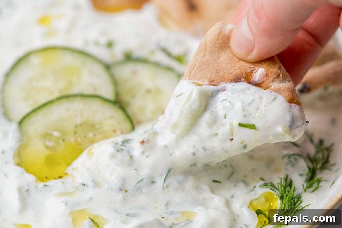A hand holds a piece of warm pita bread, perfectly dipped into a bowl of creamy Greek Tzatziki, illustrating its perfect consistency for scooping.